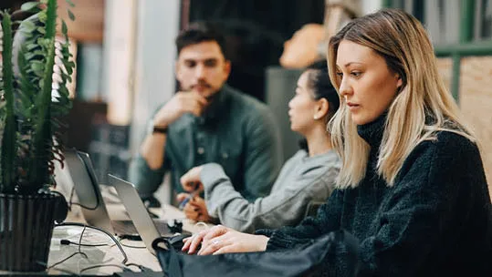 woman in office wearing black working on a laptop sitting next to two people also on laptops