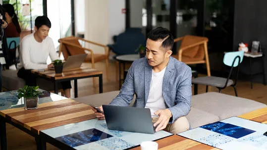 asian man at laptop on a desk looking at his mobile phone