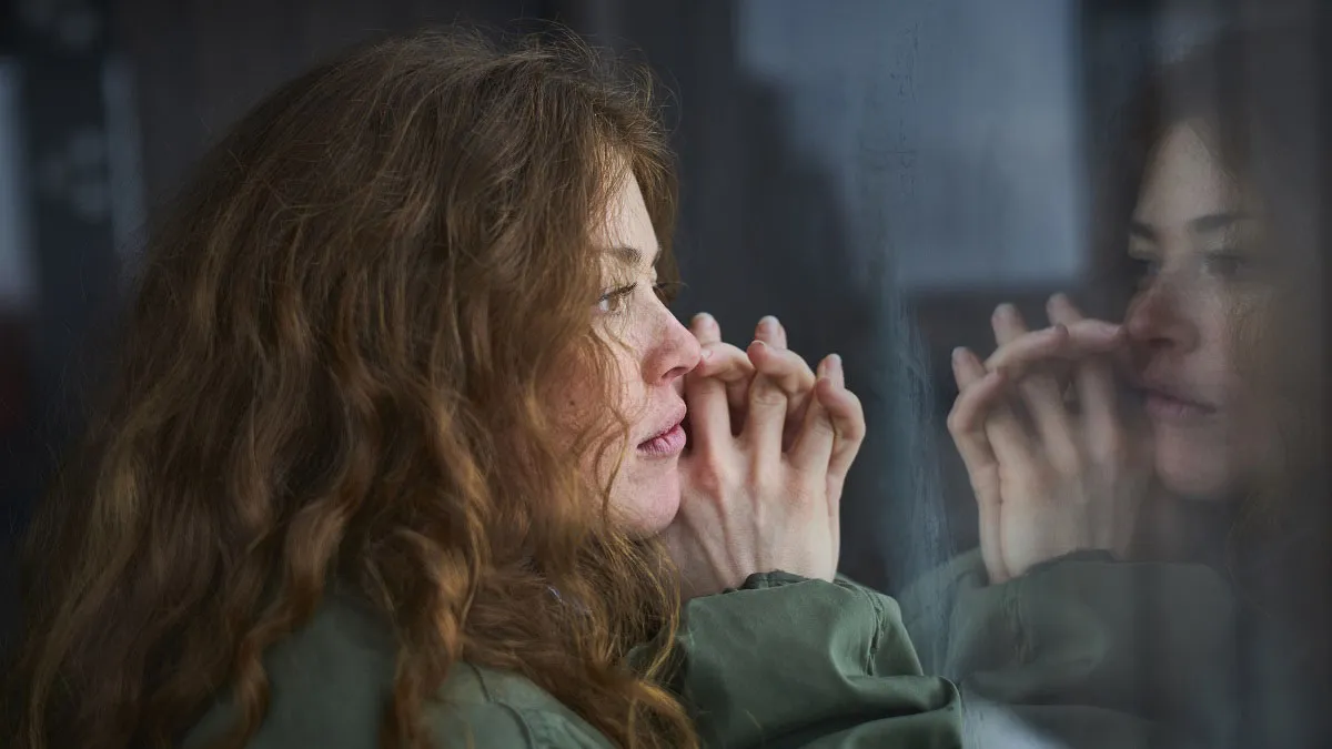 woman with curly red hair looking through a window with hands clasped