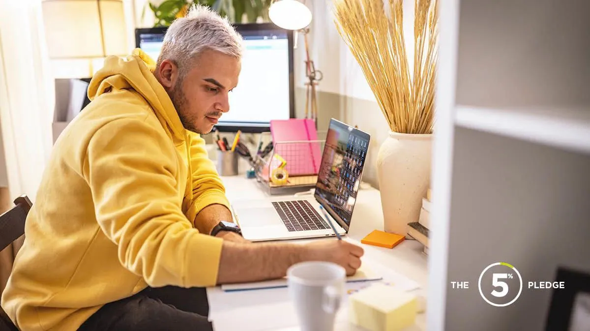 Young man working long hours at home office desk, laptop on, writing in a notepad 