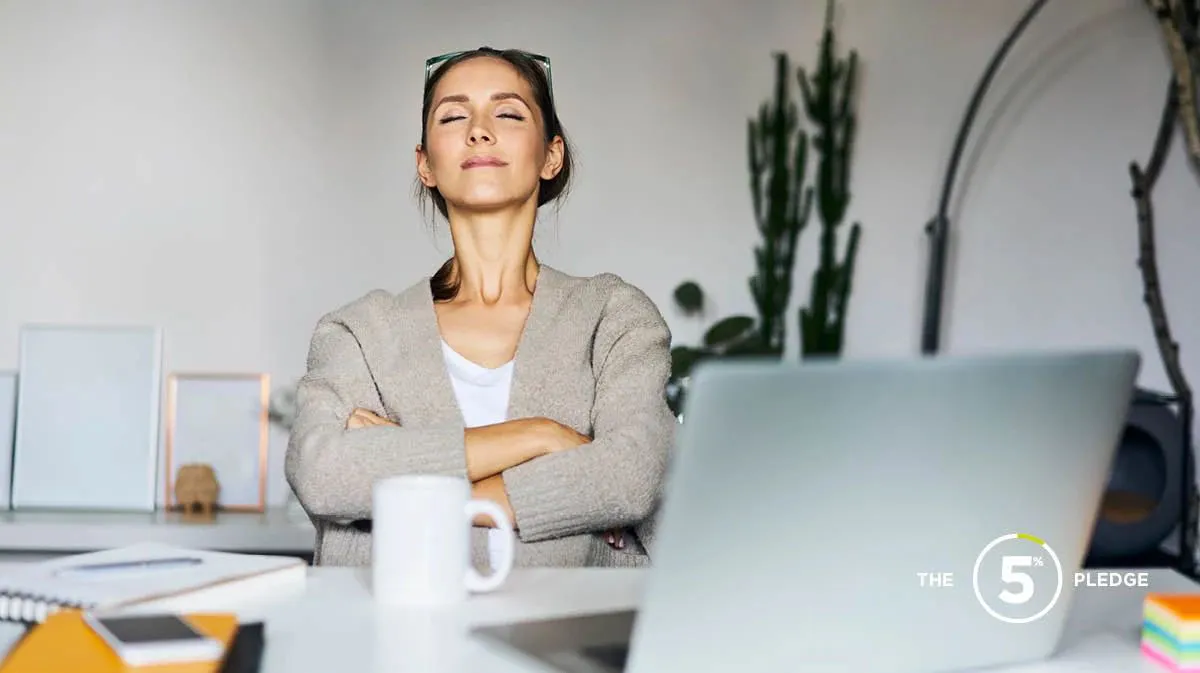 Young female sitting back in her chair at a desk in front of laptop, arms crossed, eyes closed, looking relaxed using meditat