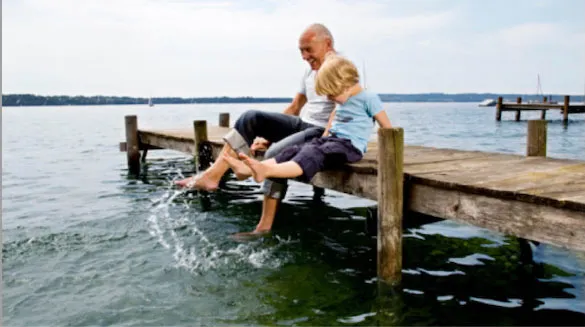 Homme âgé assis avec un enfant en bas âge sur une jetée, riant et faisant éclabousser leurs pieds dans l’eau