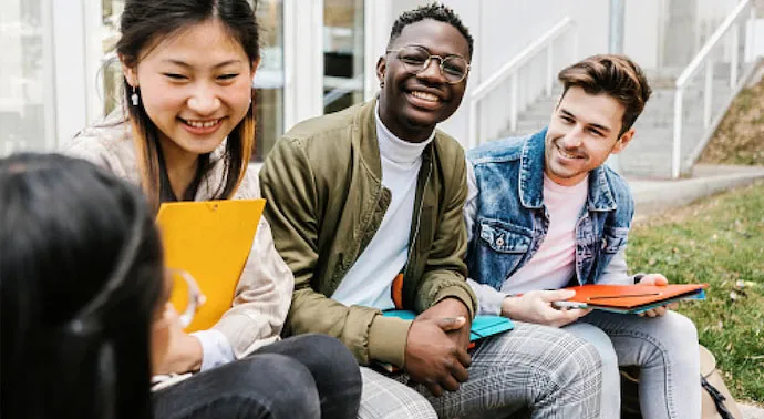 four young students sitting together holding books laughing and talking