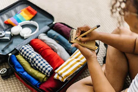 A woman sitting next to a an open suitcase filled with clothes, making notes on a paper pad