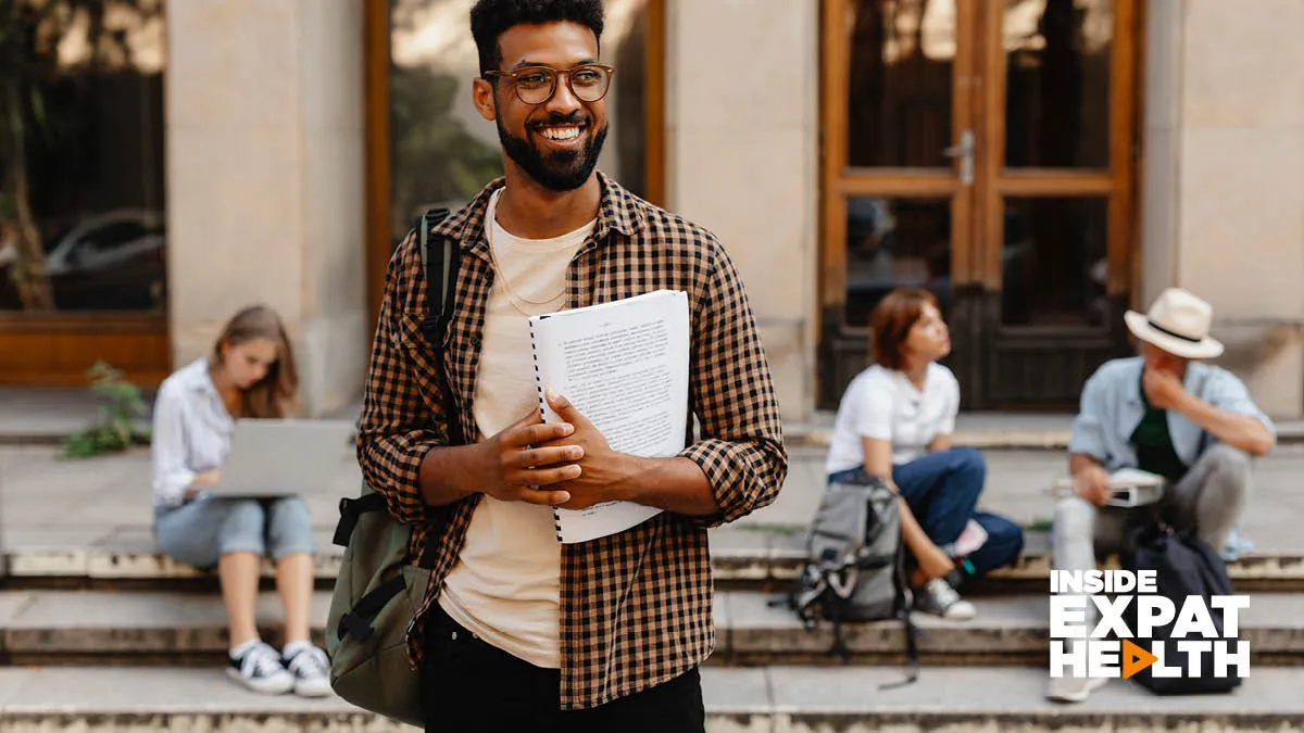 A young man smiling, holding a notebook, with students sitting on steps behind him