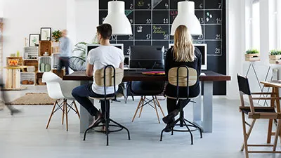the back of woman and man sitting on a large dining table in front of big computer monitors working