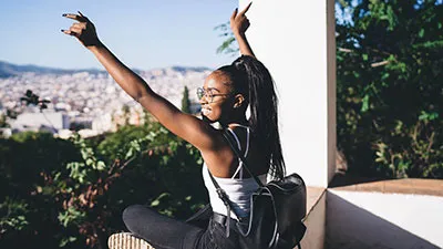 young woman wearing black backpack sitting at a viewpoint with hands in the air