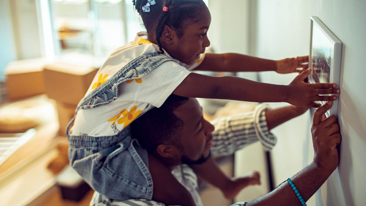 Father and daughter hanging a picture on the wall, the girl is on her father’s shoulders