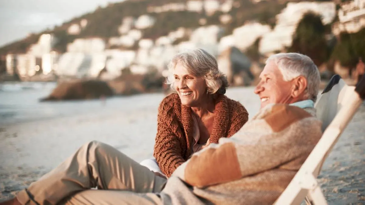 retiree couple on the beach in deck chairs laughing staring into the distance