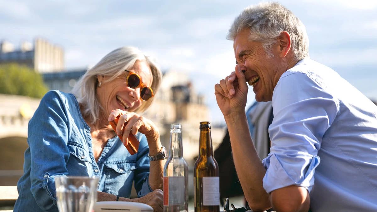 two retirees sharing a beer outdoors looking at each other laughing