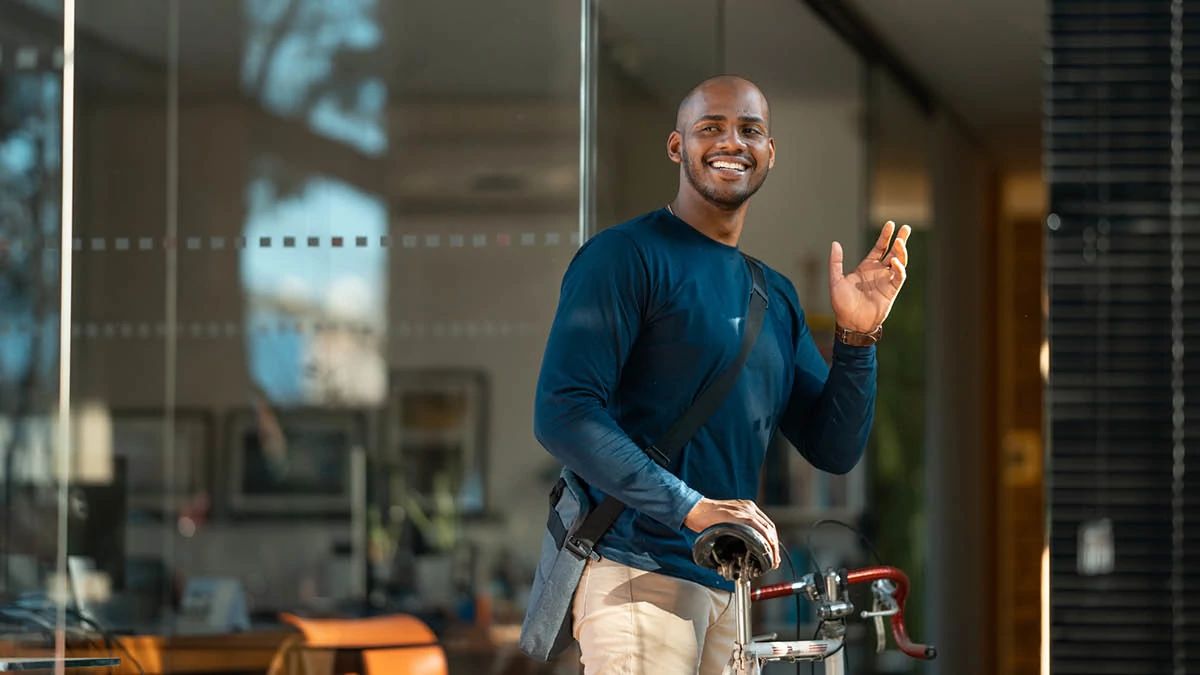 young man standing outside a glass office building, holding bike saddle and waving with his left hand