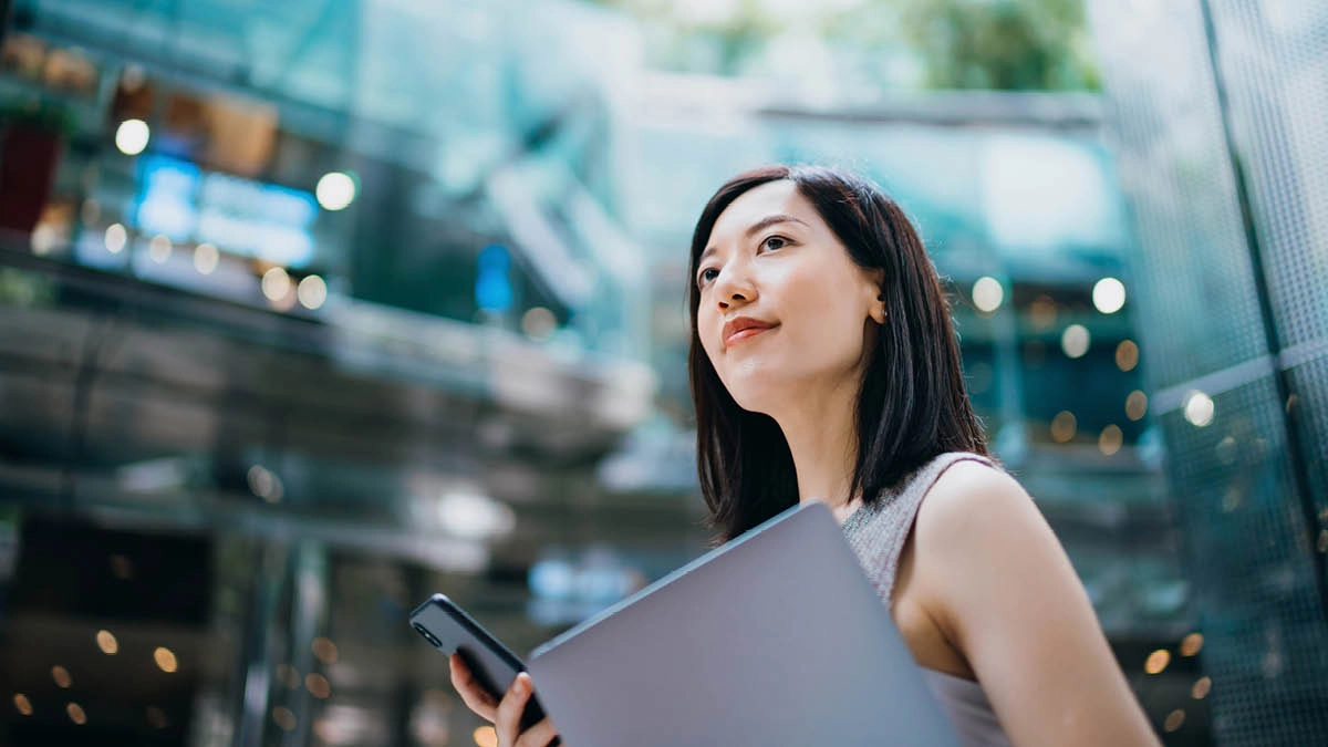 Yound Asian woman in a corporate setting, holding a laptop and mobile phone looking into the distance